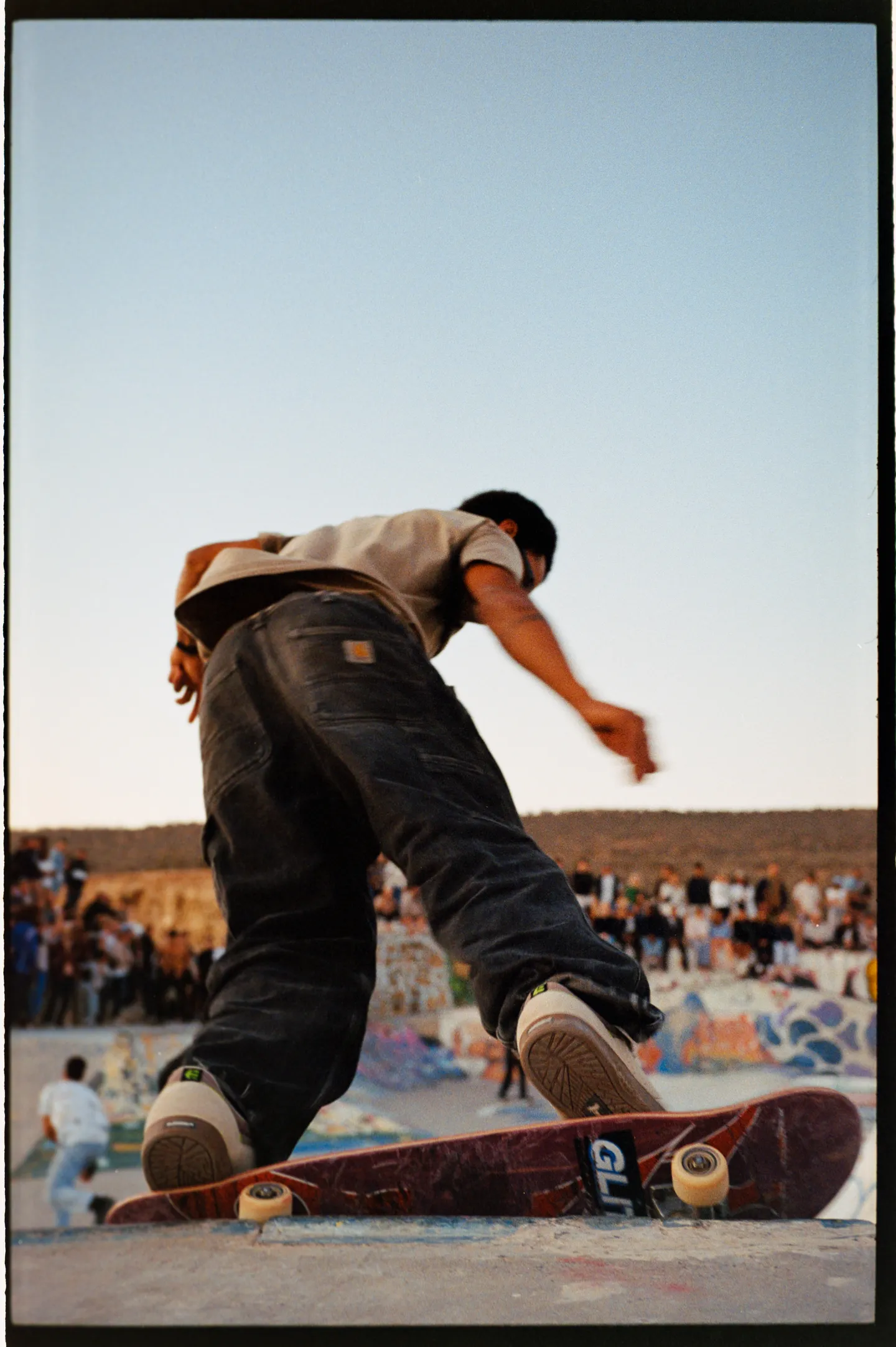 Ein Skater aus bodennaher Perspektive in Bewegung auf seinem Brett, mit Publikum am Rand des Skateparks.