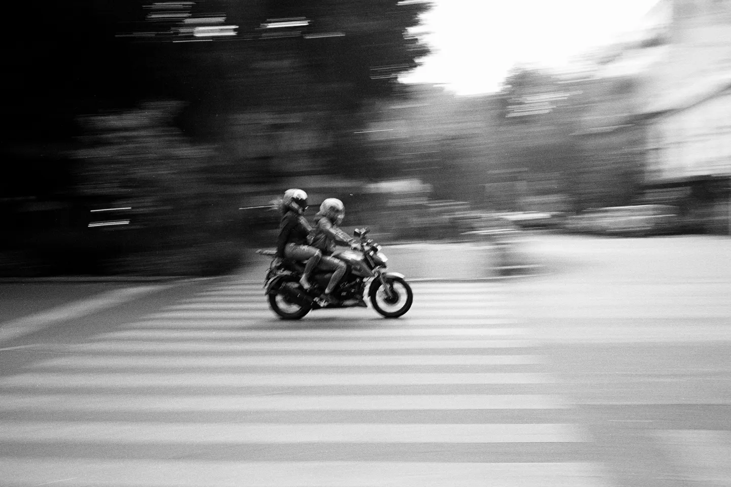Zwei Menschen auf einem Motorrad am Zebrastreifen, in Schwarz-Weiß mit mitgezogener Bewegungsunschärfe.
