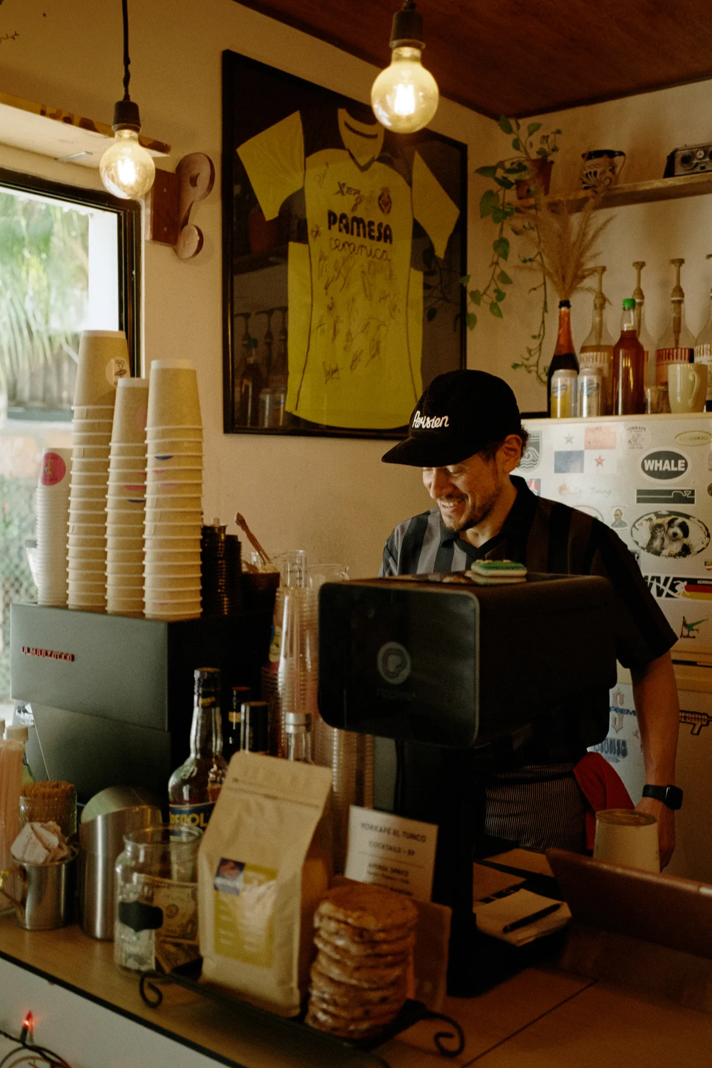 Ein lächelnder Mann hinter einer Kaffeemaschine in einem kleinen Café mit warmem Licht und hängenden Glühbirnen.