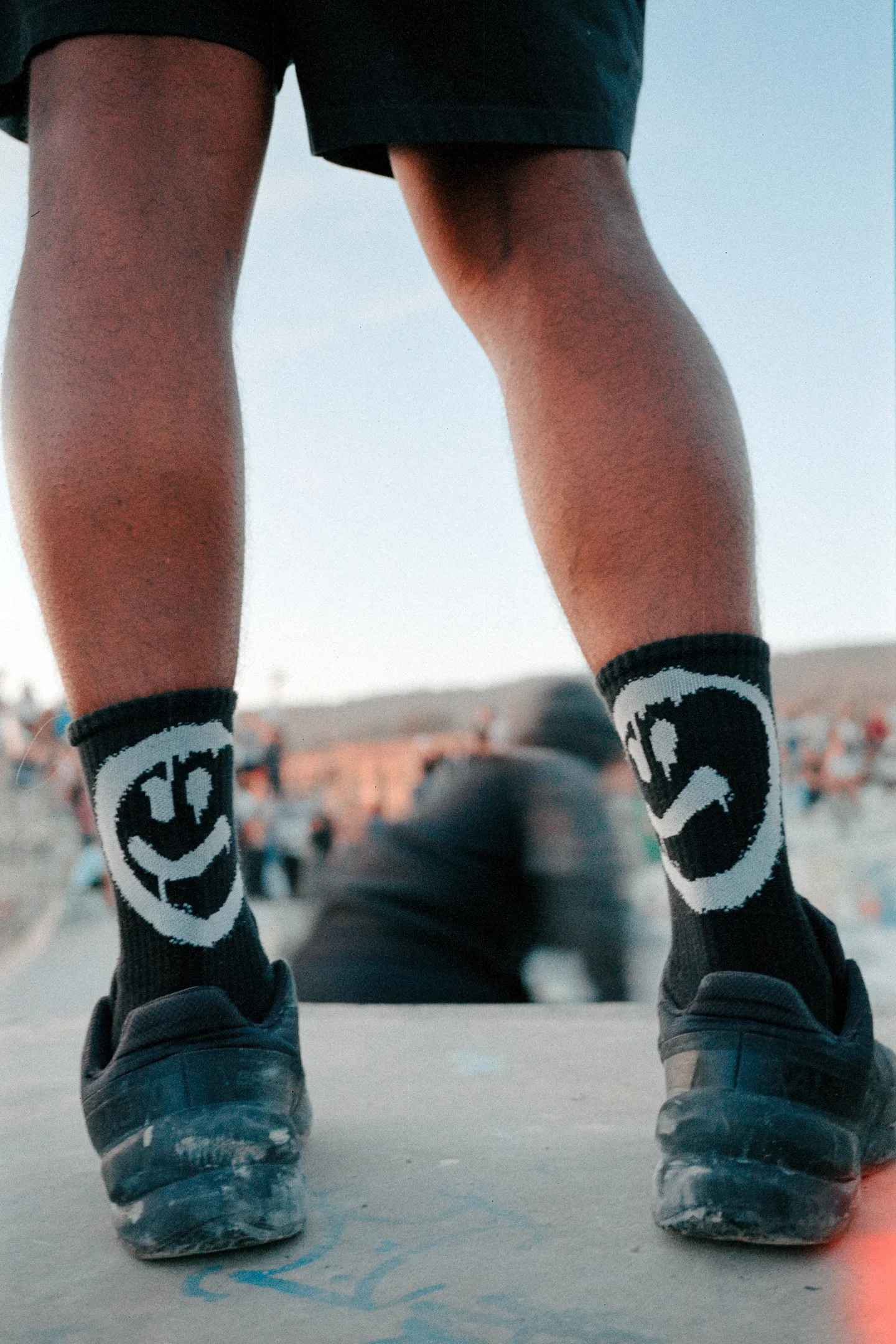 Beine eines Skaters mit Smiley-Socken auf einem Skateboard vor blauem Himmel.