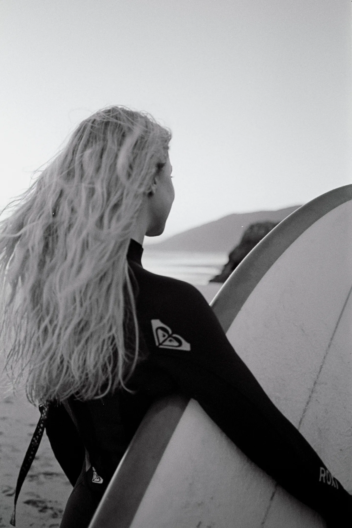 Schwarz-Weiß-Aufnahme eines Surfers mit langen Haaren und einem Surfbrett unter dem Arm am Meer.