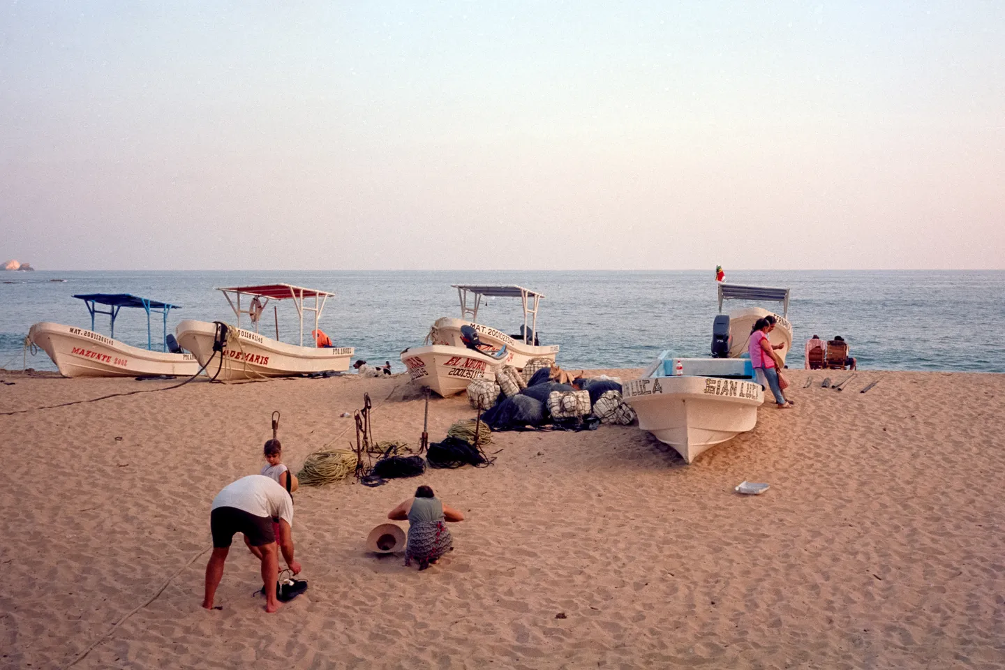 Menschen im Sand vor kleinen Fischerbooten am Meer unter einem pastellfarbenen Abendhimmel.