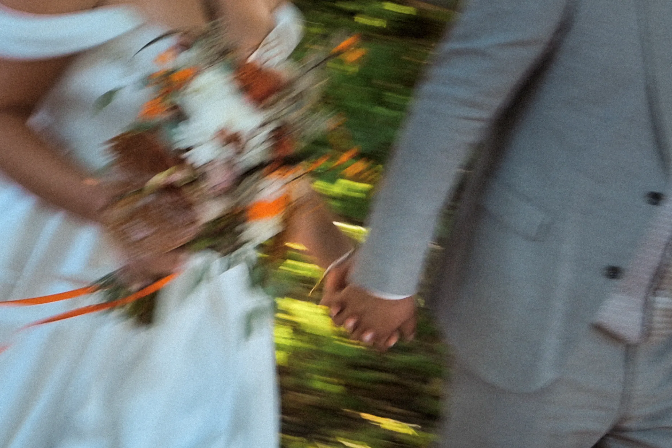 Ein Hochzeitspaar hält sich an den Händen; Brautstrauß, weißes Kleid und heller Anzug verschwimmen in der Bewegung.