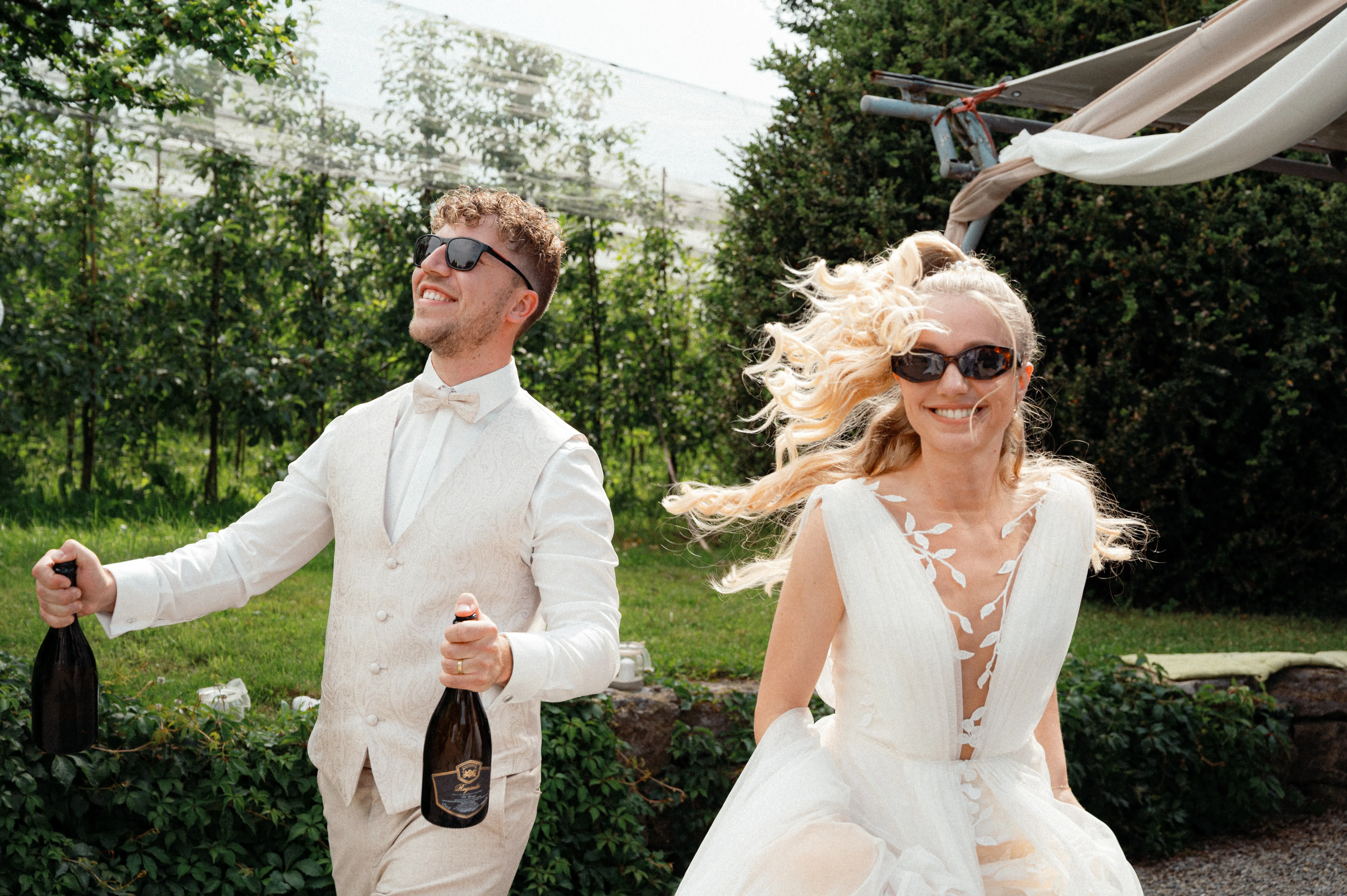 Franziska und Daniel lachen mit Sonnenbrillen im Garten; Daniel hält zwei Flaschen in den Händen.