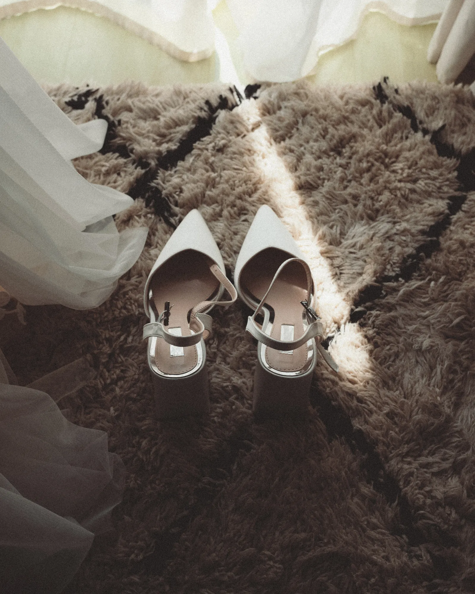 Zwei helle Brautschuhe stehen im Streiflicht auf einem Teppich, daneben fällt das Brautkleid ins Bild.