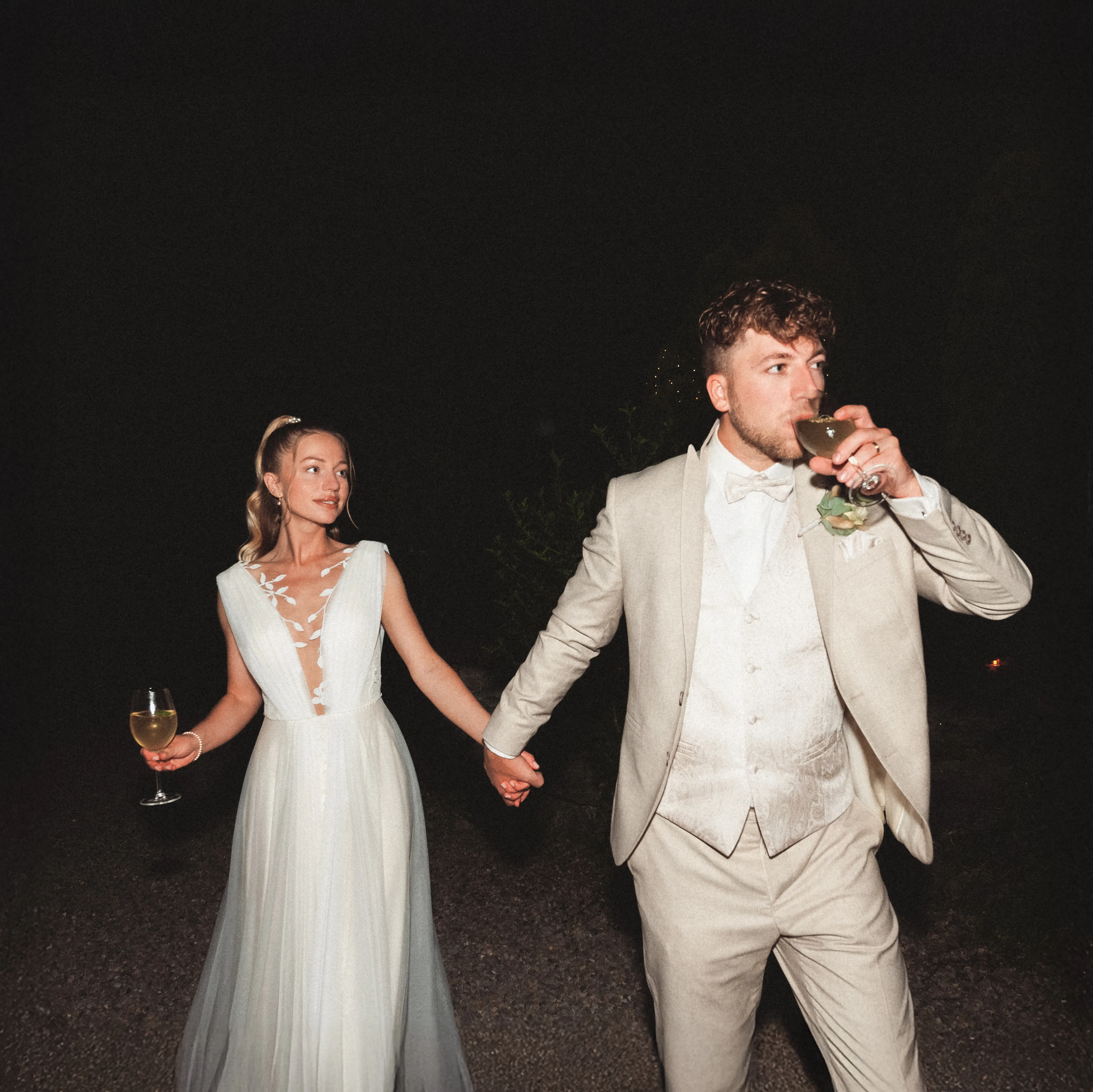 Franziska und Daniel gehen nachts Hand in Hand; beide halten ein Glas.