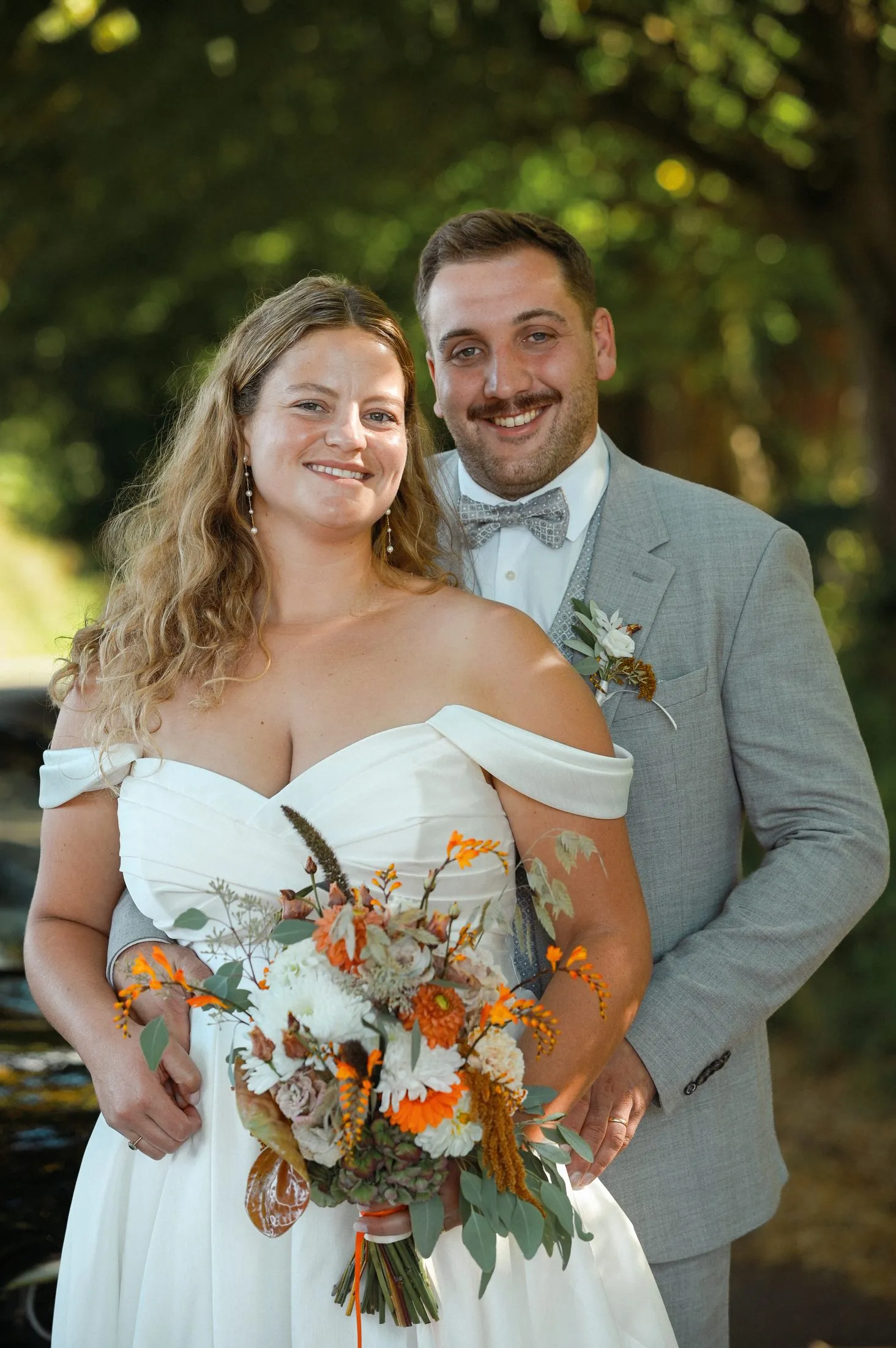 Lisa und Martin stehen mit Brautstrauß vor einem schwarzen Auto im Grünen.