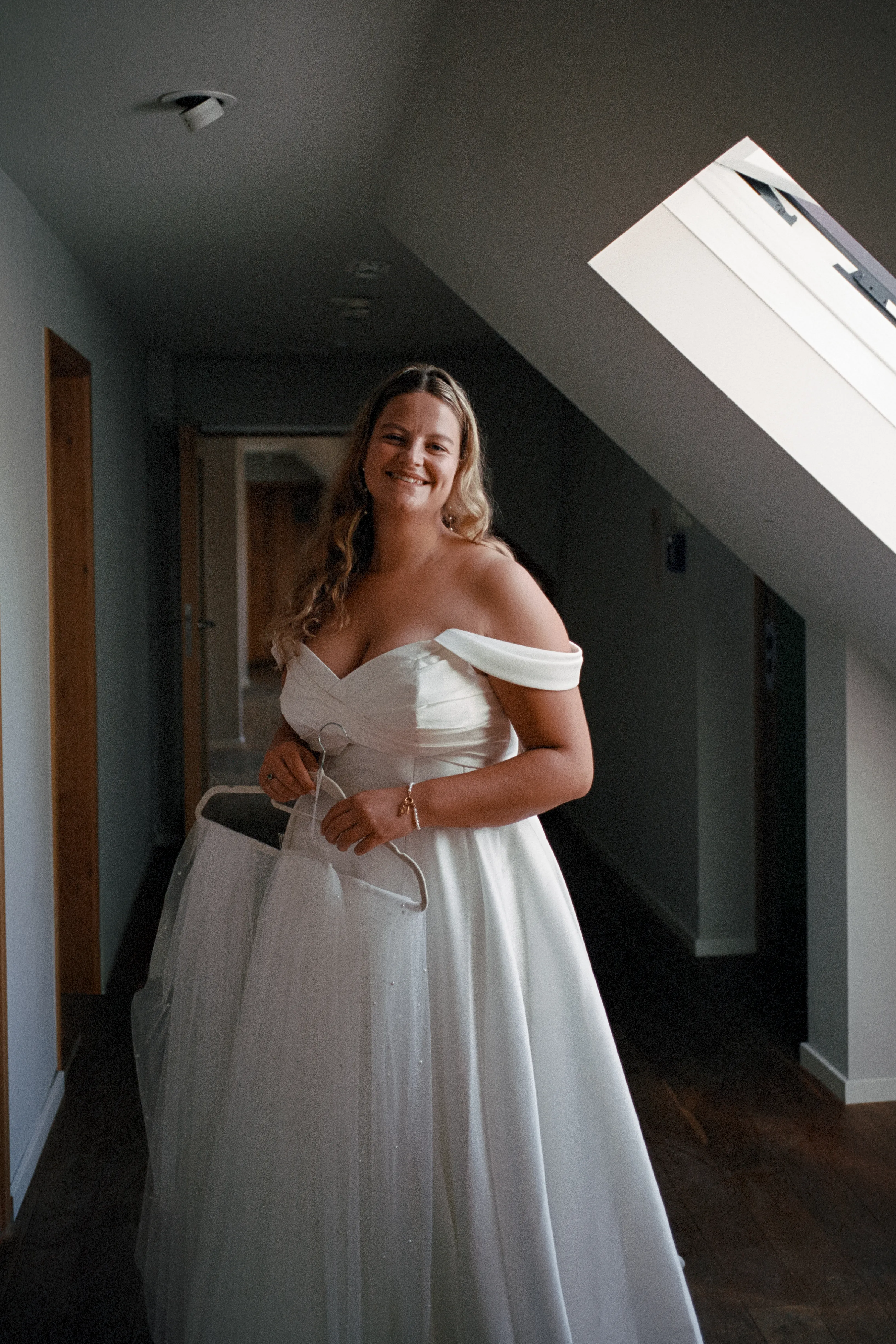 Lisa steht lächelnd in ihrem Brautkleid unter einem Dachfenster und hält ihren Schleier.