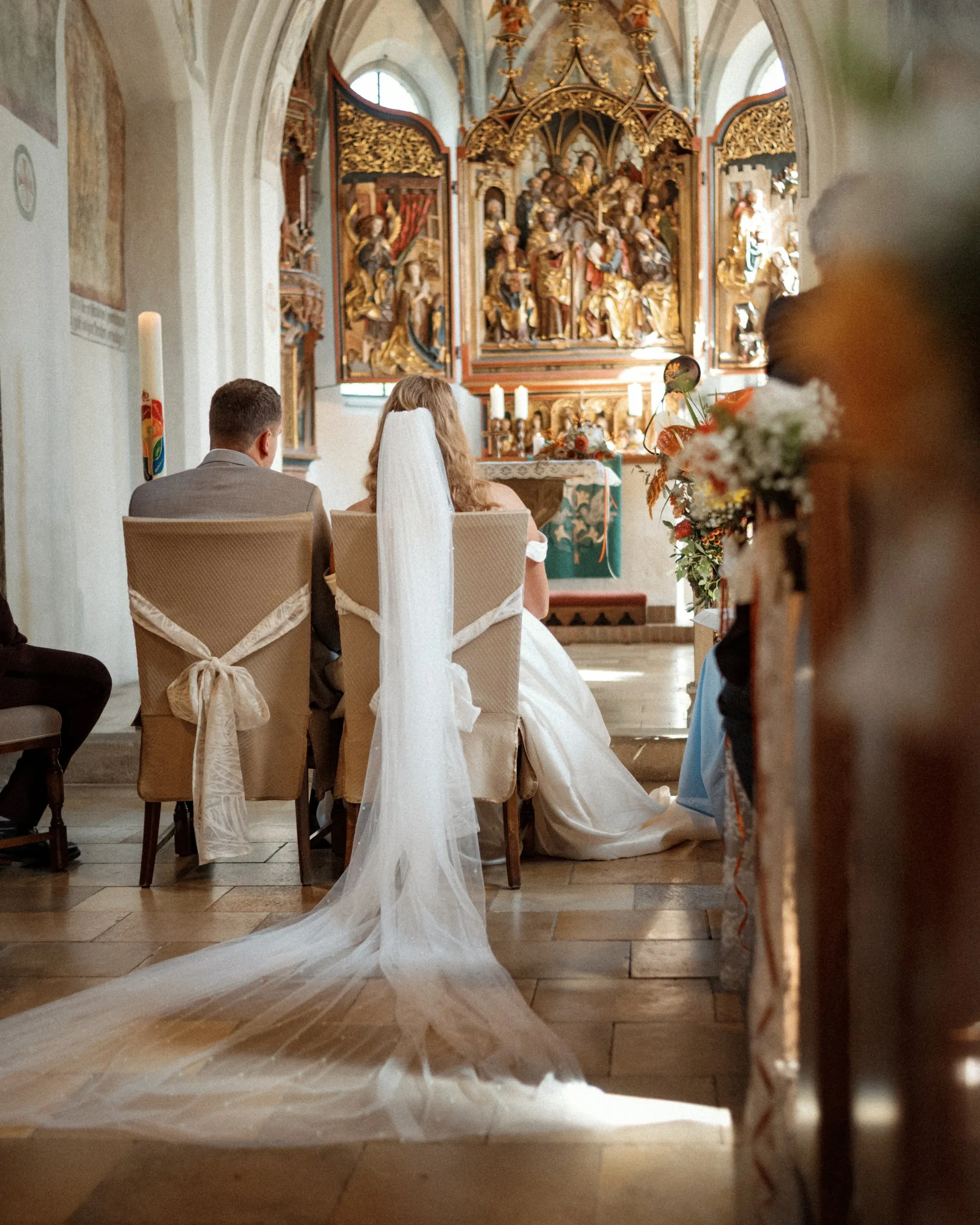 Ein Brautpaar sitzt bei der kirchlichen Trauung vor dem Altar, der Schleier der Braut liegt lang auf dem Boden.