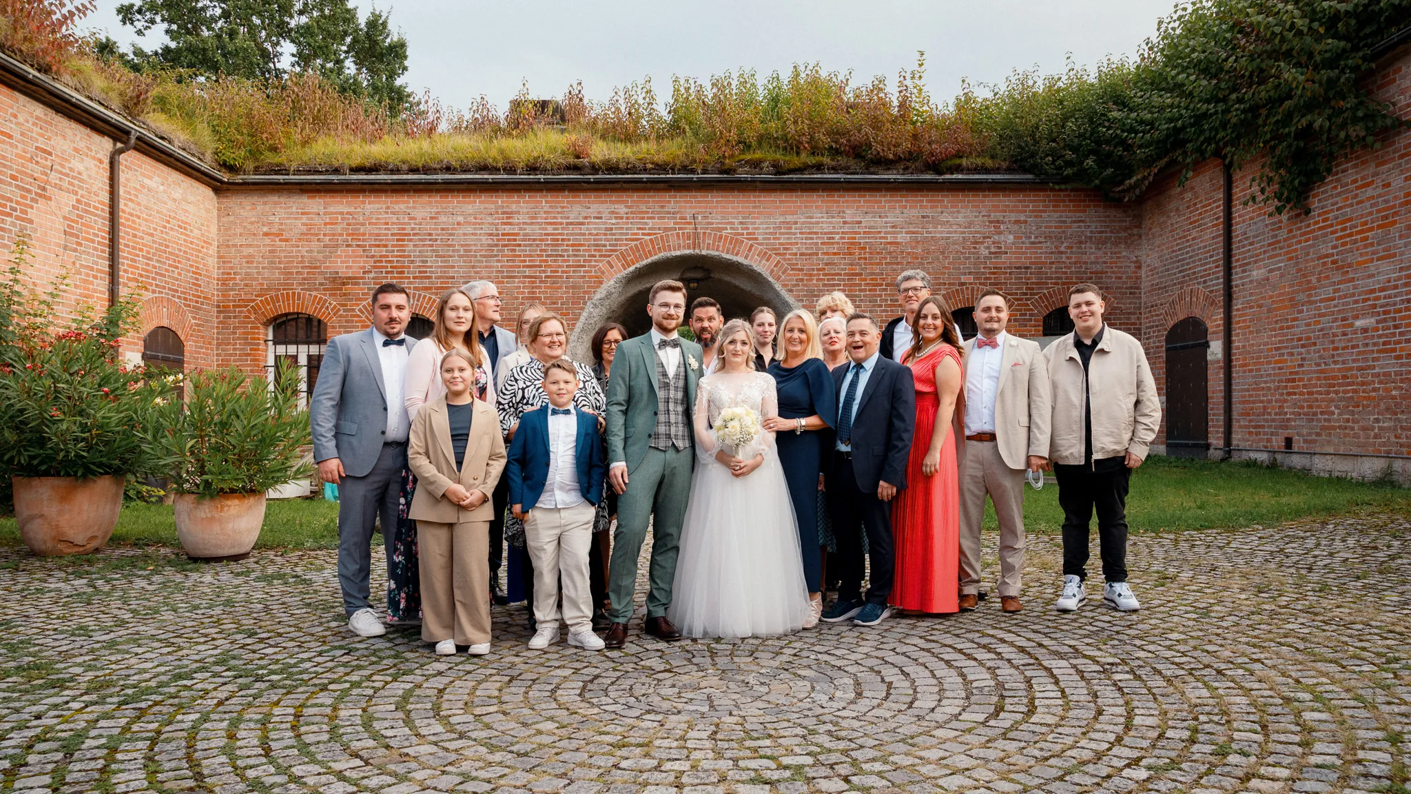 Tamara und Kevin stehen mit ihren Gästen vor einer roten Backsteinmauer.