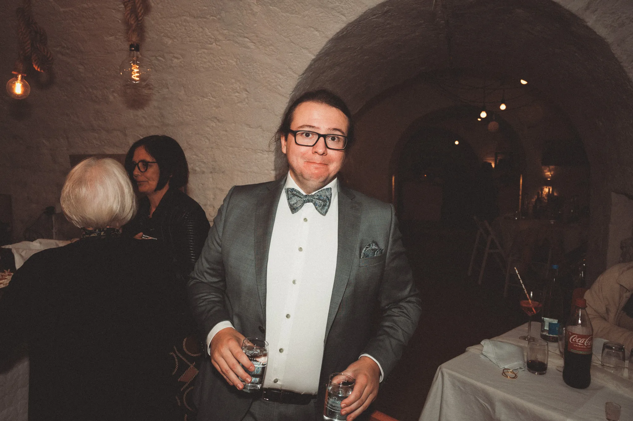 Ein Gast mit Fliege steht während der Feier mit zwei Getränken im Gewölberaum.