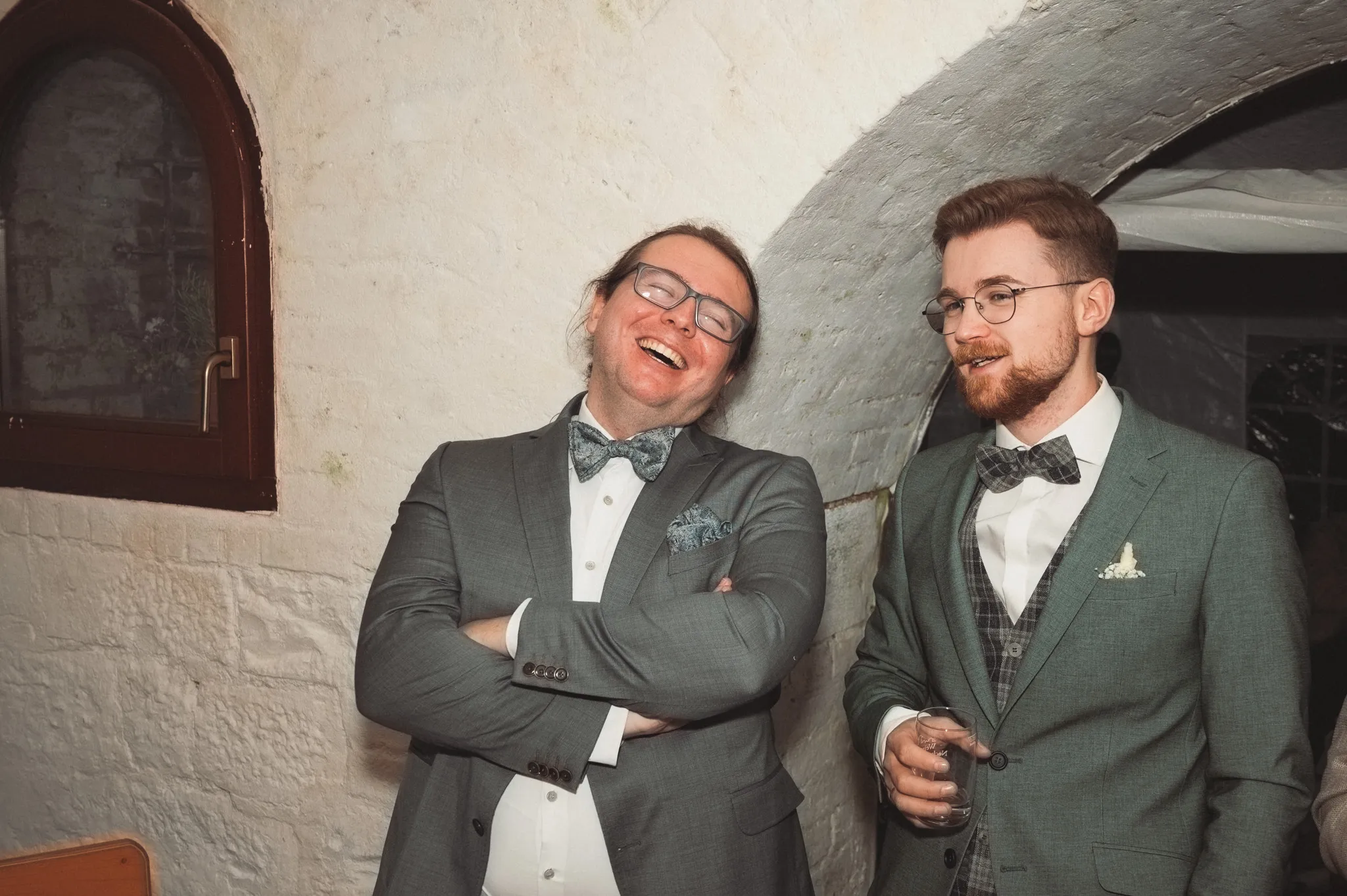 Kevin und ein Gast lachen gemeinsam an einer hellen Backsteinwand.