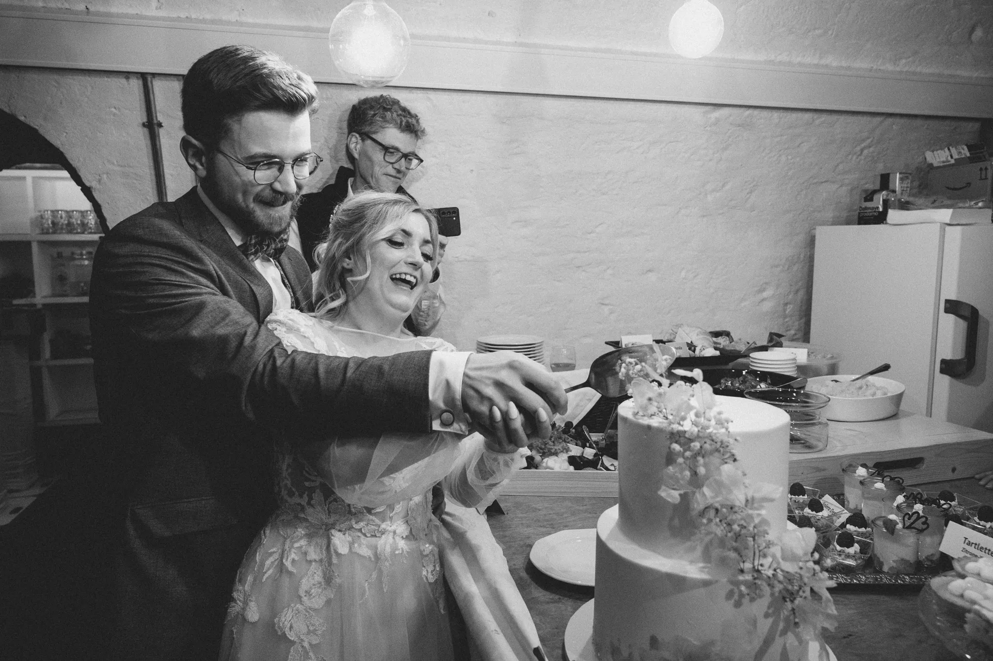 Tamara und Kevin schneiden in einer Schwarz-Weiß-Aufnahme gemeinsam die Hochzeitstorte an.