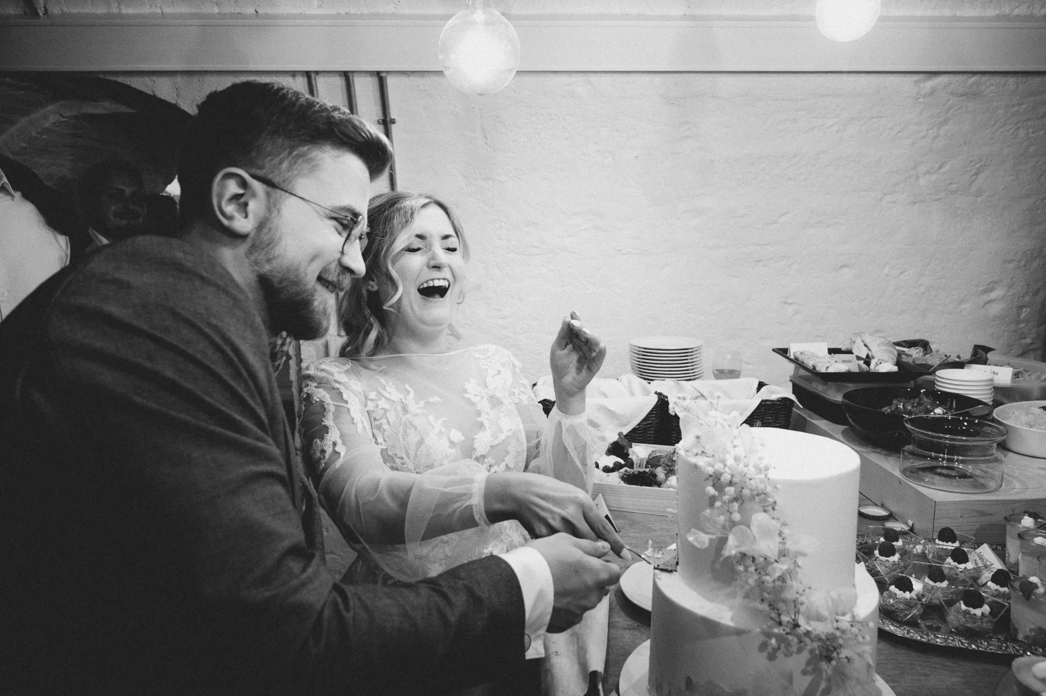 Tamara und Kevin lachen in einer Schwarz-Weiß-Aufnahme am Tisch der Hochzeitstorte.